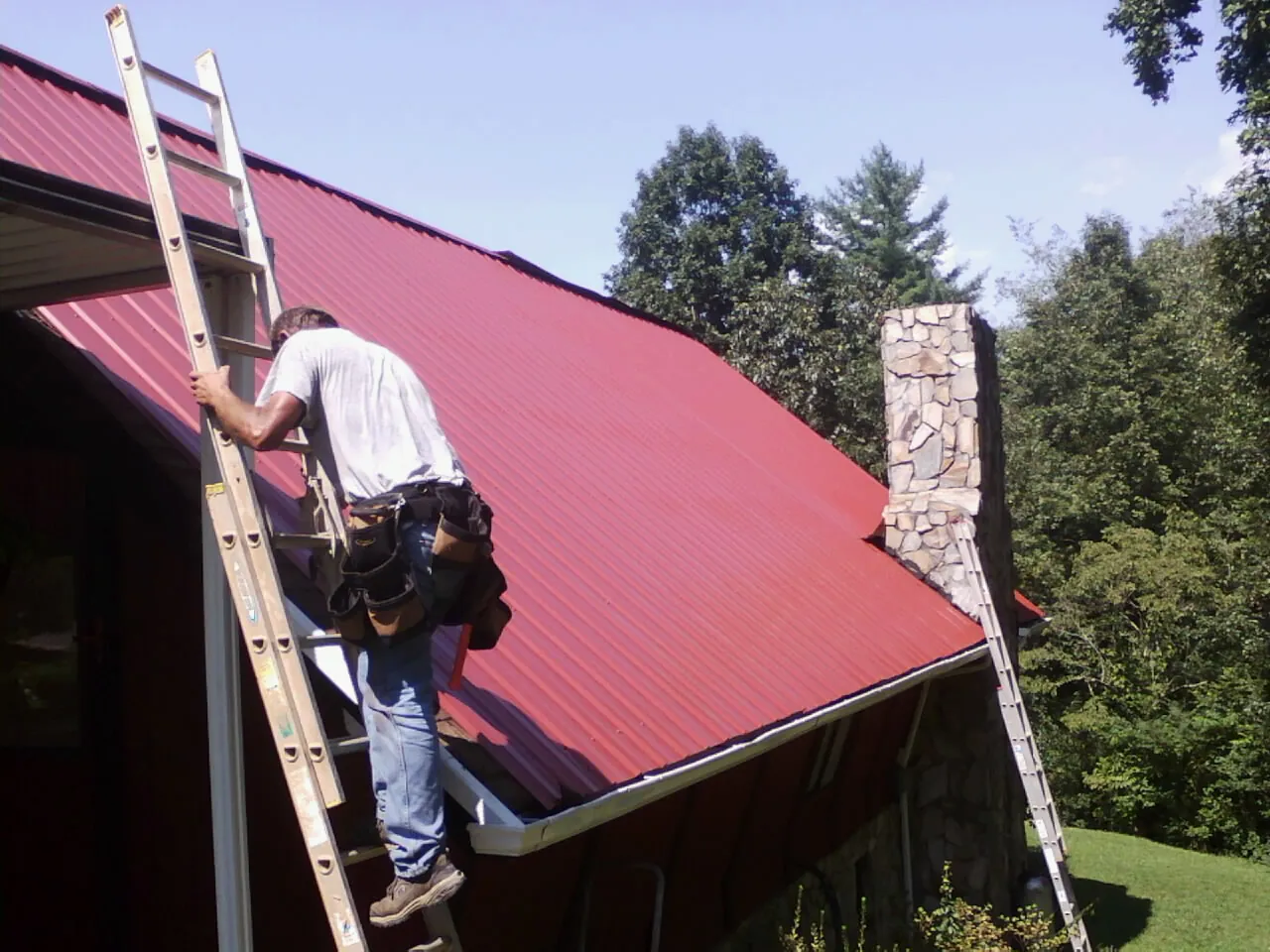 Roofer on a ladder beside a red metal roof and stone chimney — Soffit & Fascia Repair by Roofing Brothers in West Harwich
